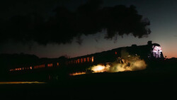 Steam locomotive at night with illuminated passenger cars and dramatic smoke plume against darkening sky.