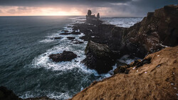 Dramatic coastal landscape with jagged rock formations and ruins silhouetted against a moody dawn sky.
