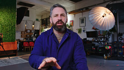 Man with beard wearing blue jacket speaks directly to camera in professional studio setting.