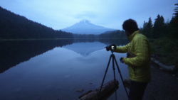 Photographer with tripod-mounted camera at dawn overlooking a calm lake with a snow-capped mountain peak reflected in the water.