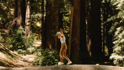 Woman in tan pants and white top jumping on a forest trail surrounded by towering redwood trees.