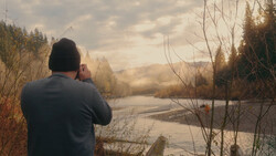 Photographer shooting a misty river landscape at golden hour with forested banks and bare branches in foreground.