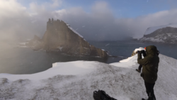 Photographer with telephoto lens on snowy ridge overlooking dramatic rocky peak and misty fjord landscape.