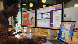 Person working at wooden desk with laptop, external monitors displaying spreadsheets and design software in modern office setting.