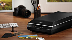 Flatbed scanner on wooden desk with photo prints, camera, and studio setup visible in background.