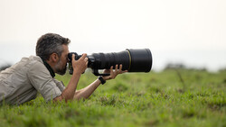 Wildlife photographer lying prone in grass, holding telephoto lens while composing a shot across an open field.