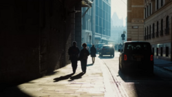 Sunlit urban street with pedestrians and parked vehicles casting long shadows on pavement.