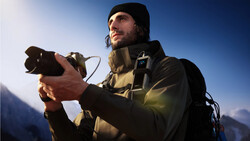 Male photographer holding a camera against a clear blue sky with mountains in the background.