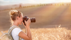 Female photographer holding a DSLR camera up to her face while standing in a golden wheat field at sunset.