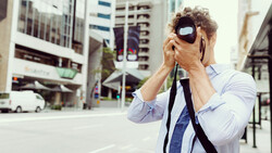 Man in light blue shirt holding camera to his face on an urban street.