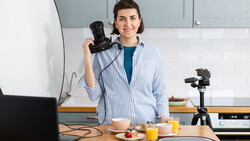 Woman holding a camera at a desk setup with a laptop, tripod-mounted camera, and food styling props.