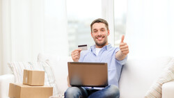 Man sitting on a couch holding a credit card and laptop while giving a thumbs up.