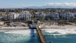 Aerial view of a wooden pier extending into turquoise ocean water with beachfront residential buildings and mountains in the background.