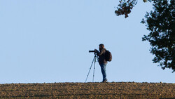 Photographer standing on a ridge silhouetted against blue sky, looking through a telephoto lens mounted on a tripod.