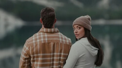 Two people standing outdoors by a lake, one wearing a tan plaid shirt facing away and one wearing a cream hoodie and beanie looking toward camera.