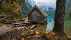 Weathered boathouse on an alpine lake shore with forested mountains and turquoise water.