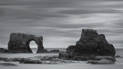 Rock formations with natural arch rising from shallow ocean water under dramatic cloudy sky.
