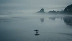 Surfer carrying board walking into misty water with forested coastline and rock formations reflected in calm sea.
