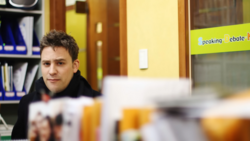 Young man in dark jacket looking at camera in bright interior space with yellow walls and shelving.