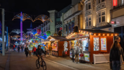 Nighttime holiday market street with illuminated vendor stalls, decorative light installations, and pedestrians shopping.