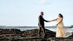 Couple holding hands on rocky beach with ocean and sky in background.