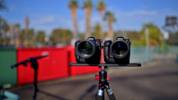 Two cinema cameras mounted on a rig against a blurred outdoor background with palm trees and red equipment.