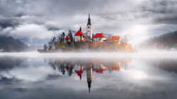 Church with red roof on misty island reflected in still water at dawn.