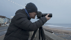 Photographer using a telephoto lens on a tripod while positioned on an elevated walkway overlooking a beach.