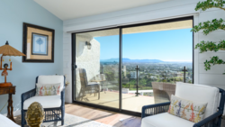 Bright bedroom with open sliding glass doors revealing a sprawling mountain valley landscape and clear blue sky.
