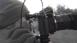 Photographer holding a camera with telephoto lens up to their eye, viewed from the side profile outdoors.
