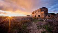 Abandoned stone building at sunset with golden light rays streaming across overgrown fields.