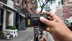 Hand holding a rangefinder film camera with yellow accent on the body, mounted on a tripod in an urban street setting.