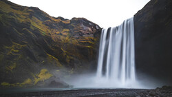 Waterfall cascading down dark basalt cliffs with moss-covered volcanic rock and mist rising from the pool below.