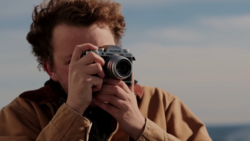 Person holding a rangefinder film camera up to their face, photographing against a coastal backdrop.