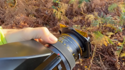 Photographer's hand holding a wide-angle lens with filter holder attached over fern-covered forest floor.