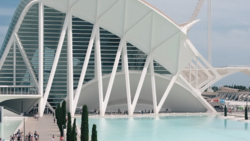 Modern white architectural structure with curved ribbed exterior beside turquoise reflecting pool, cypress trees, and glass building facade.