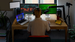 Person seated at wooden desk viewing multiple monitors displaying game graphics and code, with laptop, microphone, and lighting equipment visible.