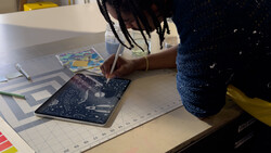Person drawing on a dark blue patterned surface with a pen while seated at a white desk with a ruler and other materials nearby.
