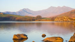 Mountain lake at golden hour with foreground rocks and tree-lined shoreline under clear sky.
