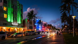 Art Deco hotel facades illuminated with neon green and blue lighting along a palm-lined street at dusk.