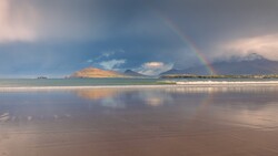 Rainbow stretching across a bay toward mountains reflected in wet sand at golden hour.