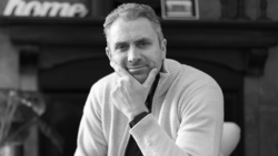 Portrait of a man in a light cardigan and dark tie against a blurred indoor background.