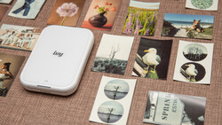 Portable photo printer surrounded by printed photos and contact sheets on a textured surface.