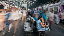 Woman sitting on a bench at a train station, surrounded by blurred commuters and purple trains.
