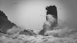 Dramatic black and white seascape with towering rock formation engulfed in crashing waves and mist.