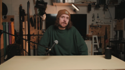 Man speaking into microphone at table in workshop with tools and lighting equipment in background.