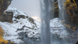 Tall waterfall cascading down snow-covered rocky cliffs with golden lichen visible on the stone.