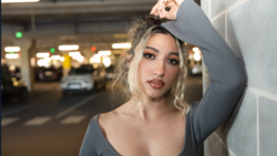 Woman with blonde hair posing against a white wall in a parking garage setting.