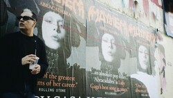 Man in sunglasses and black shirt standing in front of large promotional posters with black and white portrait imagery and text.