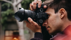 Male photographer holding a mirrorless camera to his face while focusing on a subject.